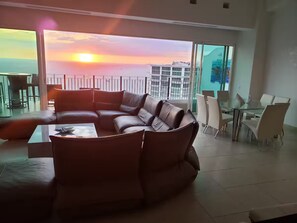 A modern living room with a large leather sofa, a glass coffee table, and a dining area with a glass table and chairs. - Beautiful beachfront penthouse with private pool (Puerto Vallarta)