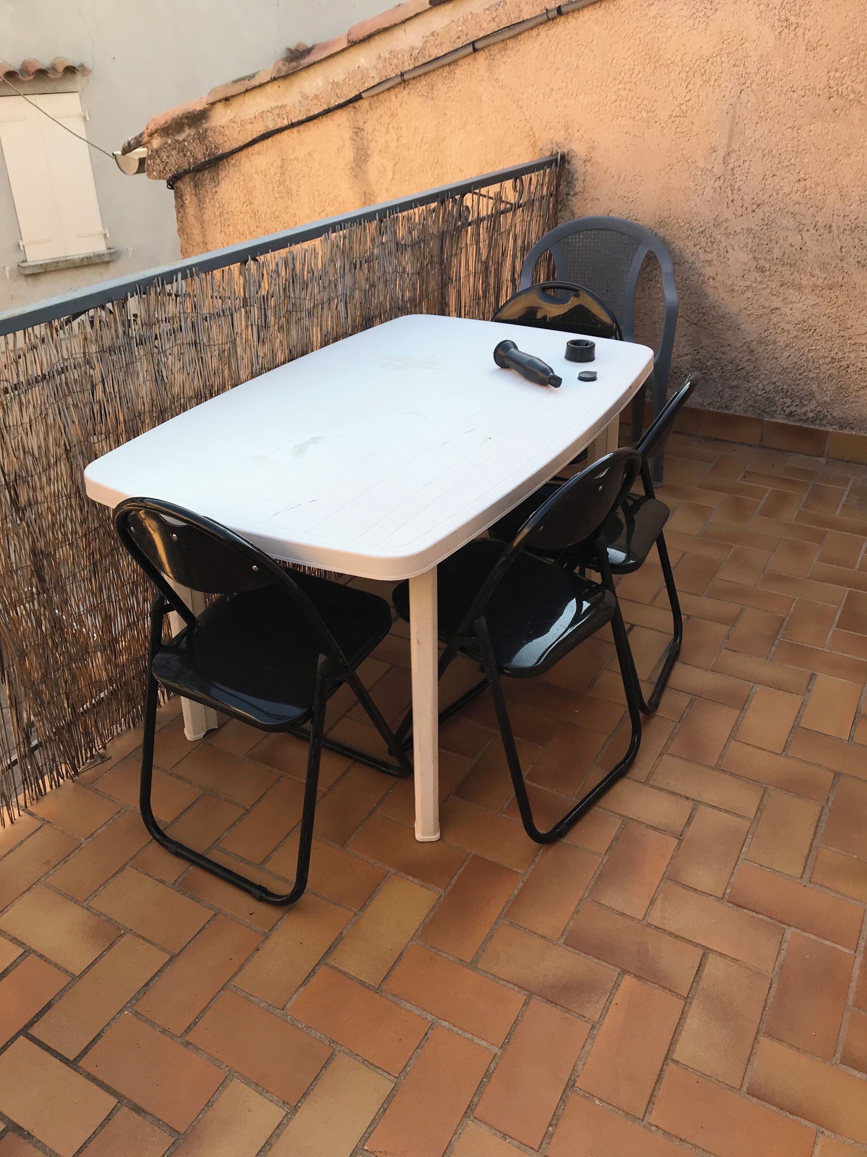 A white table with two black chairs, a bottle, and a cup on a tiled patio.