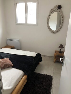 Ein Schlafzimmer mit einem Bett, einem Nachttisch, einer Lampe, einem Spiegel und einem Fenster.
