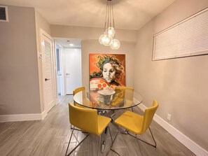 Un coin repas avec une table en verre, des chaises jaunes, un luminaire au plafond et un tableau accroché au mur.
