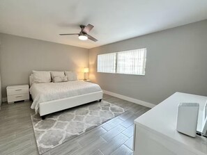 Une chambre à coucher avec un lit, une table de chevet, une lampe, une fenêtre et un ventilateur de plafond.