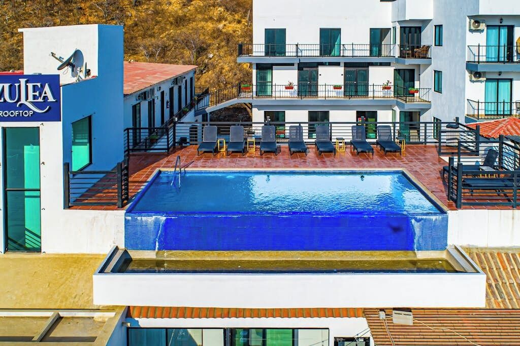 A rooftop pool with a modern building in the background, a sign for 'ZUEA ROOFTOP,' and outdoor seating.