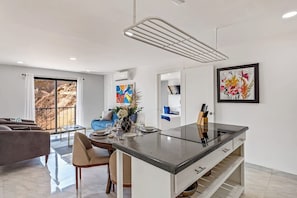 Una cucina moderna con isola centrale, zona pranzo con tavolo e sedie, e un soggiorno con divano e un'ampia finestra.