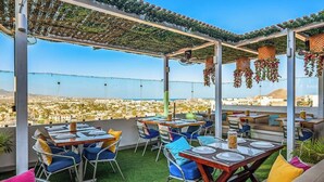 An outdoor dining area with tables and chairs, a pergola with hanging decorations, and a view of a cityscape. - Morgan Residences 508 @ Boutique Hotel With Pool, Gym & Spa (Cabo San Lucas)