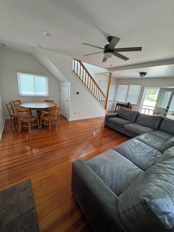 A spacious living area with a wooden floor, a gray sofa, a dining table with chairs, and a staircase.
