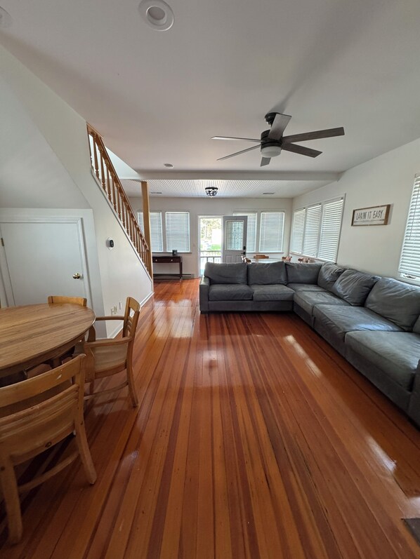 A living room with a wooden floor, a gray sectional sofa, a wooden dining table with chairs, a ceiling fan, and a staircase.