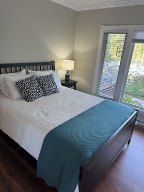 A bedroom with a bed, pillows, a bedside table with a lamp, a window with blinds, and a view of greenery outside. - Suite 3 for the win! Secluded suite overlooking the harbour, Spectacular views (Thetis Island)