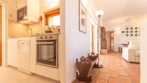 Uma cozinha compacta com micro-ondas, forno e lava-louças, um corredor com decoração em formato de coroa, e uma sala de estar com sofá branco.