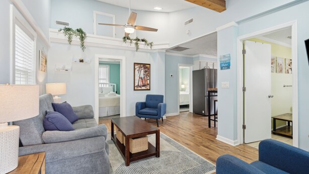 A spacious living room with a ceiling fan, a blue armchair, a grey sofa, and a wooden coffee table. - Sunny Side Up - Heart of Folly Two Bedroom Stay (Folly Beach)