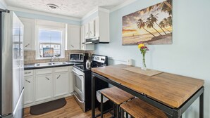 A kitchen with white cabinets, a wooden table, and a painting of a beach scene. - Sunny Side Up - Heart of Folly Two Bedroom Stay (Folly Beach)
