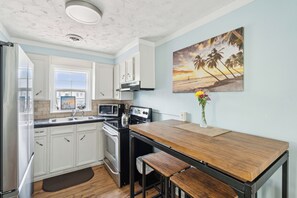 A kitchen with white cabinets, a wooden table, and a painting of a beach scene. - Sunny Side Up - Heart of Folly Two Bedroom Stay (Folly Beach)