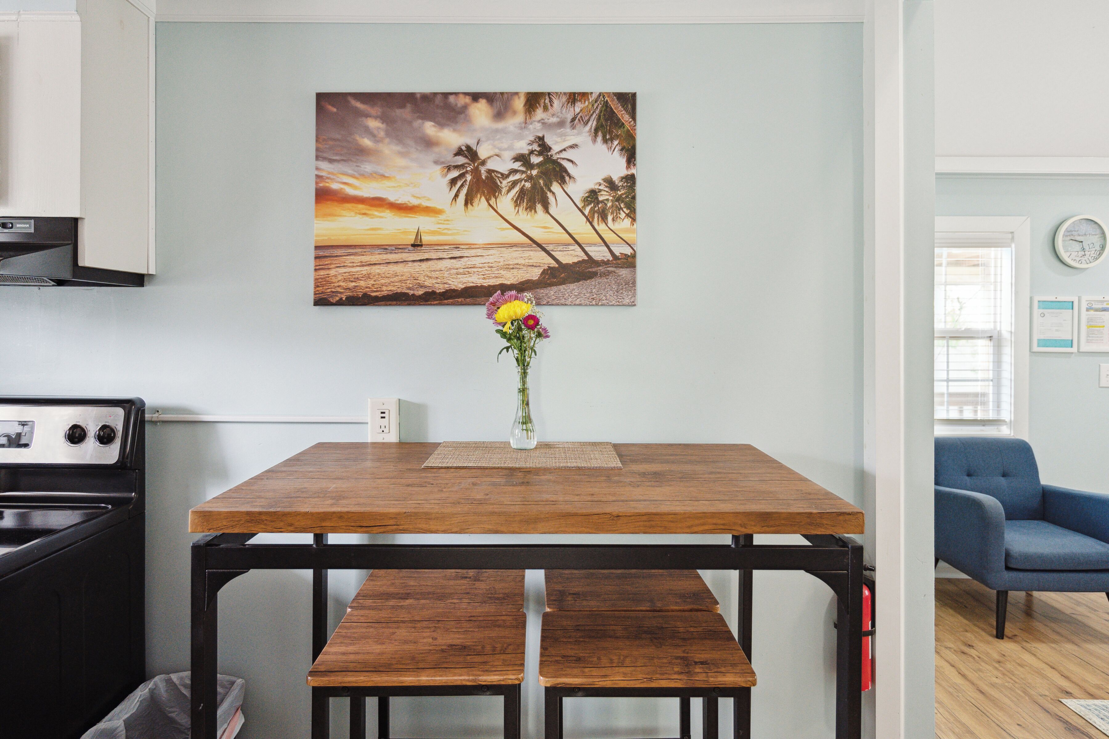 Una cocina con estufa, una mesa de comedor con bancos, un reloj en la pared y un cuadro con una escena de playa.