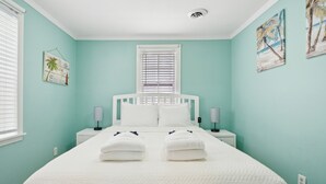 A bedroom with a bed, two bedside tables, a window with blinds, and wall art. - Sunny Side Up - Heart of Folly Two Bedroom Stay (Folly Beach)