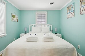 A bedroom with a bed, two bedside tables, a window with blinds, and wall art. - Sunny Side Up - Heart of Folly Two Bedroom Stay (Folly Beach)