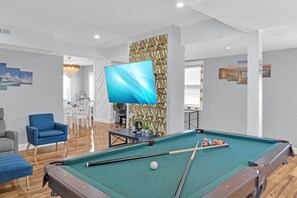 A pool table with a green felt surface, a blue armchair, and a white dining table with chairs in a room with wooden flooring and a decorative wall panel. - Perfect 4-bedroom house with EV Charger in pleasant Buffalo neighborhood (Buffalo)