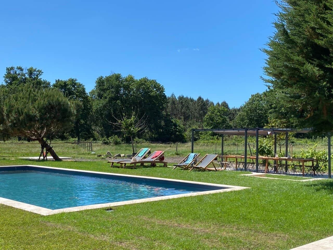 Une piscine rectangulaire entourée d’une pelouse, un espace de repos couvert avec des tables, et des arbres en arrière-plan.
