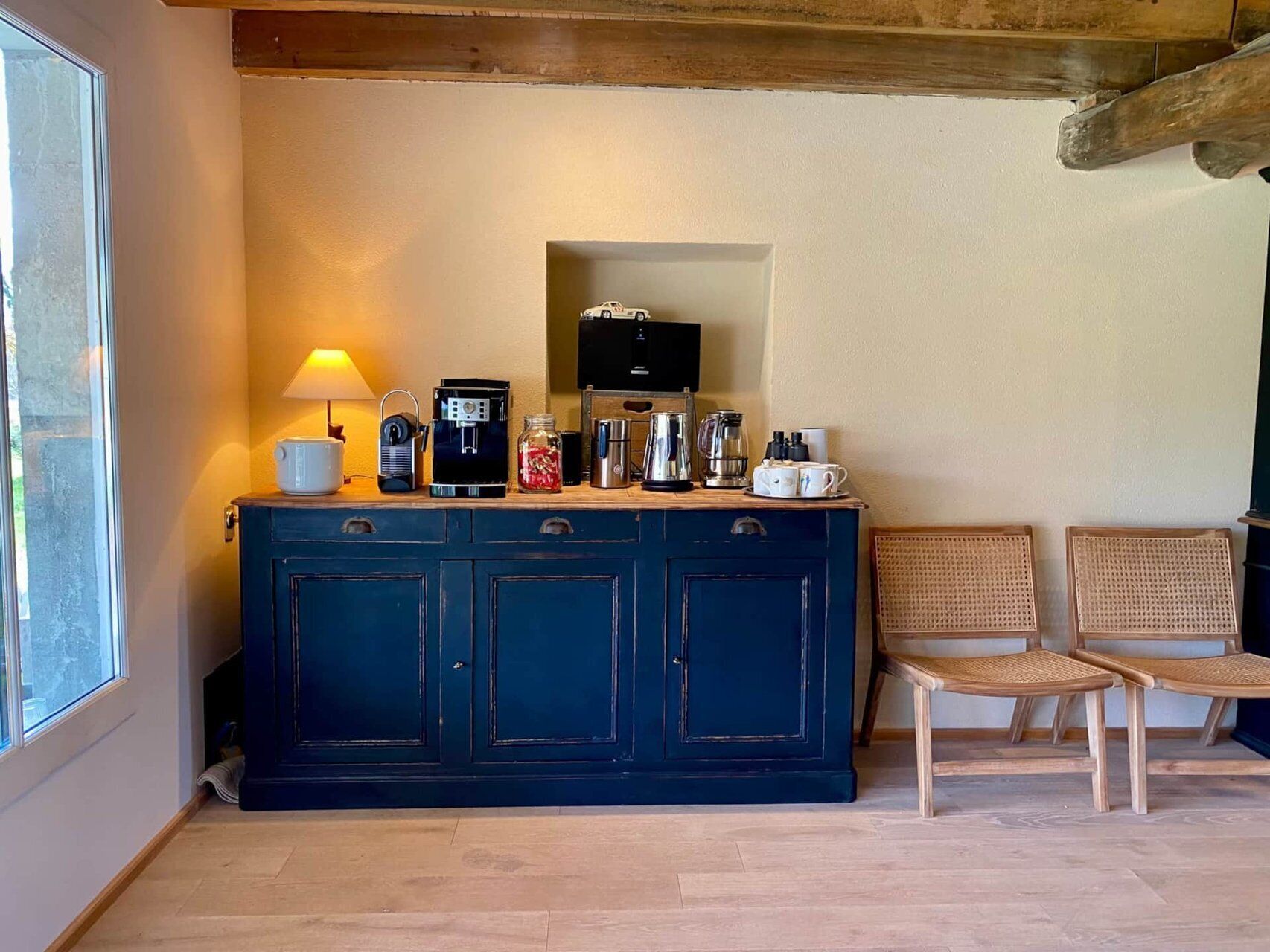 A kitchenette with a coffee maker, a kettle, and a coffee pot on a cabinet. There are two wicker chairs and a window to the left.