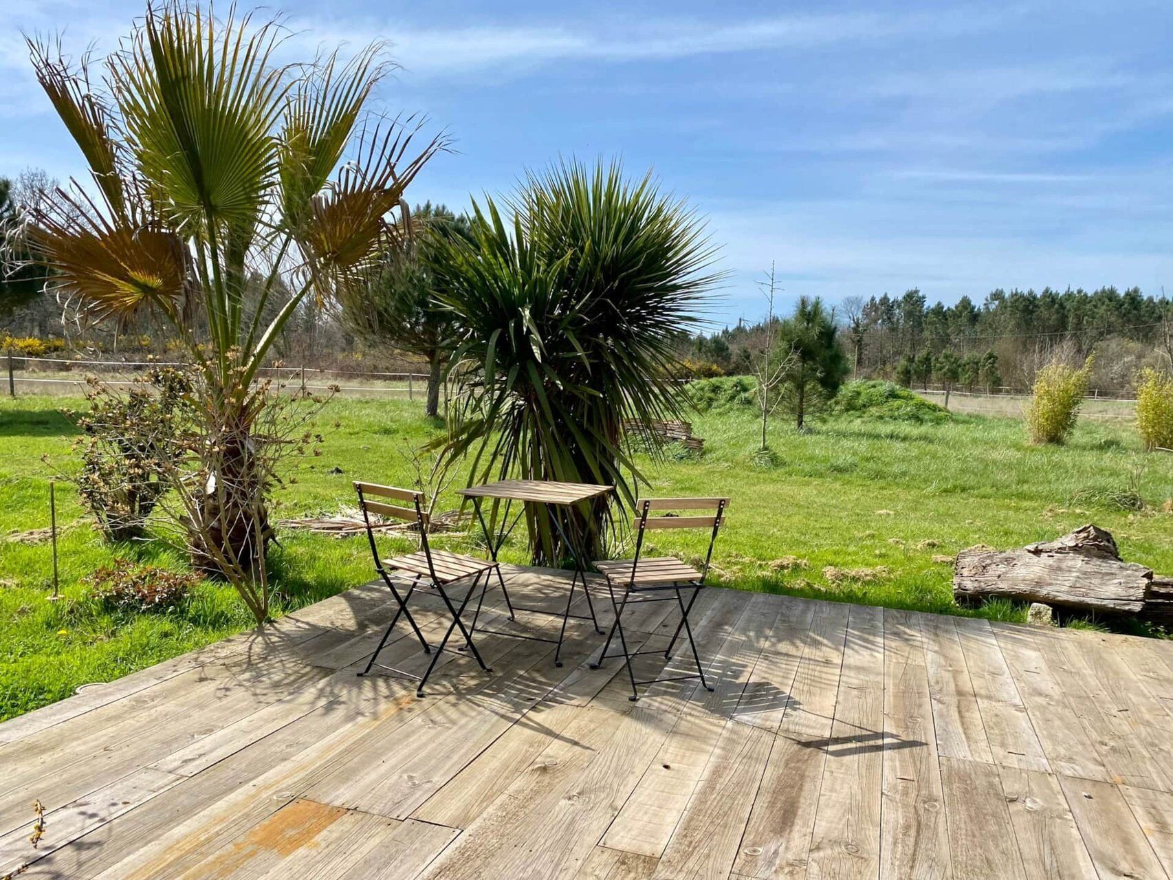 Eine Holzterrasse im Freien mit zwei Holzstühlen und einem Tisch, umgeben von viel Grün und blauem Himmel.