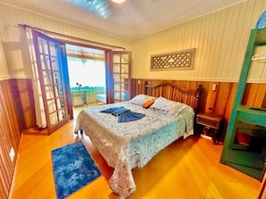 A bedroom with a bed, a wooden headboard, a nightstand, a window with curtains, and a view of the outdoors.