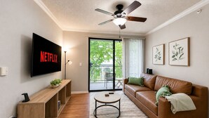 Un salon moderne doté d’un téléviseur à écran plat, d’un ventilateur de plafond, d’une console en bois, d’un canapé en cuir et d’un balcon offrant une vue sur la verdure.
