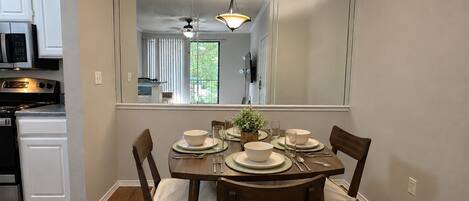 A dining area with a table set for four, a kitchen in the background, and a large mirror reflecting the room.