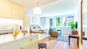Una habitación de hotel moderna con kitchenette, una cama con ropa de cama azul, un sofá, un televisor y un ventanal con vistas a zonas verdes.