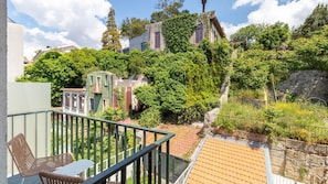 A balcony with a wicker chair and table, overlooking a garden with trees and a stone wall. - Central & Trendy w/ Balcony by LovelyStay (Porto)