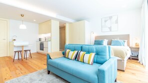 Moderni hotellihuone, jossa on sininen sohva, pieni keittotila, erillinen ruokapöytä ja sängyssä pääty.