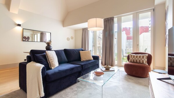 Ett modernt vardagsrum med en mörkblå soffa, ett soffbord i glas och en röd fåtölj. Det finns ett stort fönster med gardiner, en platt-TV och en spegel på väggen.