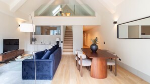 Ein modernes Interieur mit einer Schlafgalerie, einem Essbereich mit Holztisch und -stühlen, einem dunkelblauen Sofa und einem Fernseher auf einem Holzgestell.