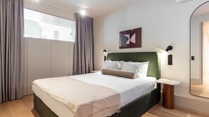 Ett modernt hotellrum med en stor säng, ett sängbord, en spegel och ett fönster med gardiner.