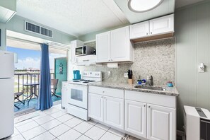 A kitchen with white cabinets, a microwave, stove, and a sink. There is a balcony with a blue curtain and a view of the outside. - Surf Studio (Corpus Christi)