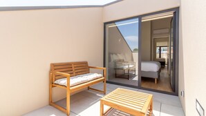 Un balcone con una panca e un tavolo di legno, e una porta scorrevole in vetro che dà su una camera da letto con un letto e una sedia.