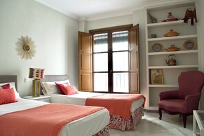 Ein Hotelzimmer mit zwei Betten, einem Sessel, einem Regal mit Dekoration und einem Fenster mit Rollläden.