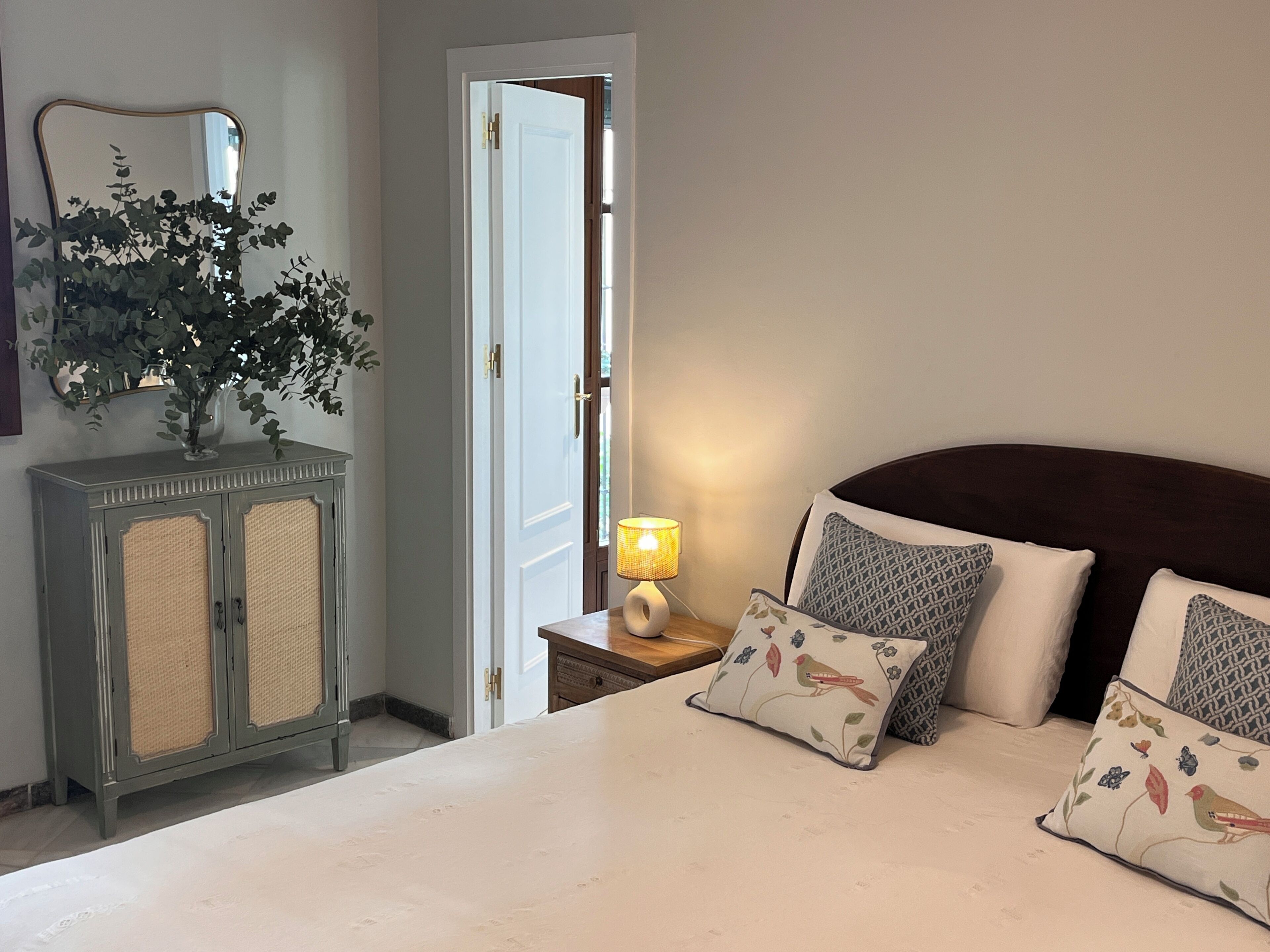 Une chambre à coucher avec un lit, des oreillers, une table de chevet avec une lampe, une commode, un miroir et une porte.