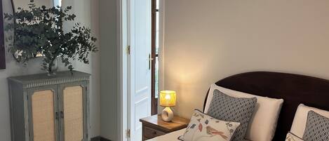 Un dormitorio con cama, almohadas, mesita de noche con lámpara, cómoda, espejo y puerta.