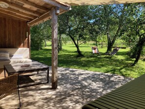 Una terraza de madera con un sofá y una mesa, rodeada de césped, árboles y una hamaca.