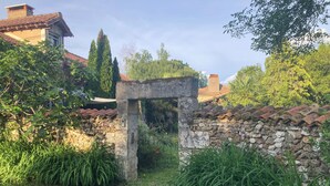 A stone archway leading to a garden area with a stone wall, surrounded by greenery and a building with a tiled roof in the background. - Atypical gite in Dordogne-Périgord, quiet, with swimming pool and beautiful view (Manzac-sur-Vern)