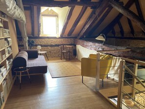 A room with a loft bed, a sofa, a small table, and a chair. There is a bookshelf filled with books, a window with a view, and a stone wall. - Atypical gite in Dordogne-Périgord, quiet, with swimming pool and beautiful view (Manzac-sur-Vern)