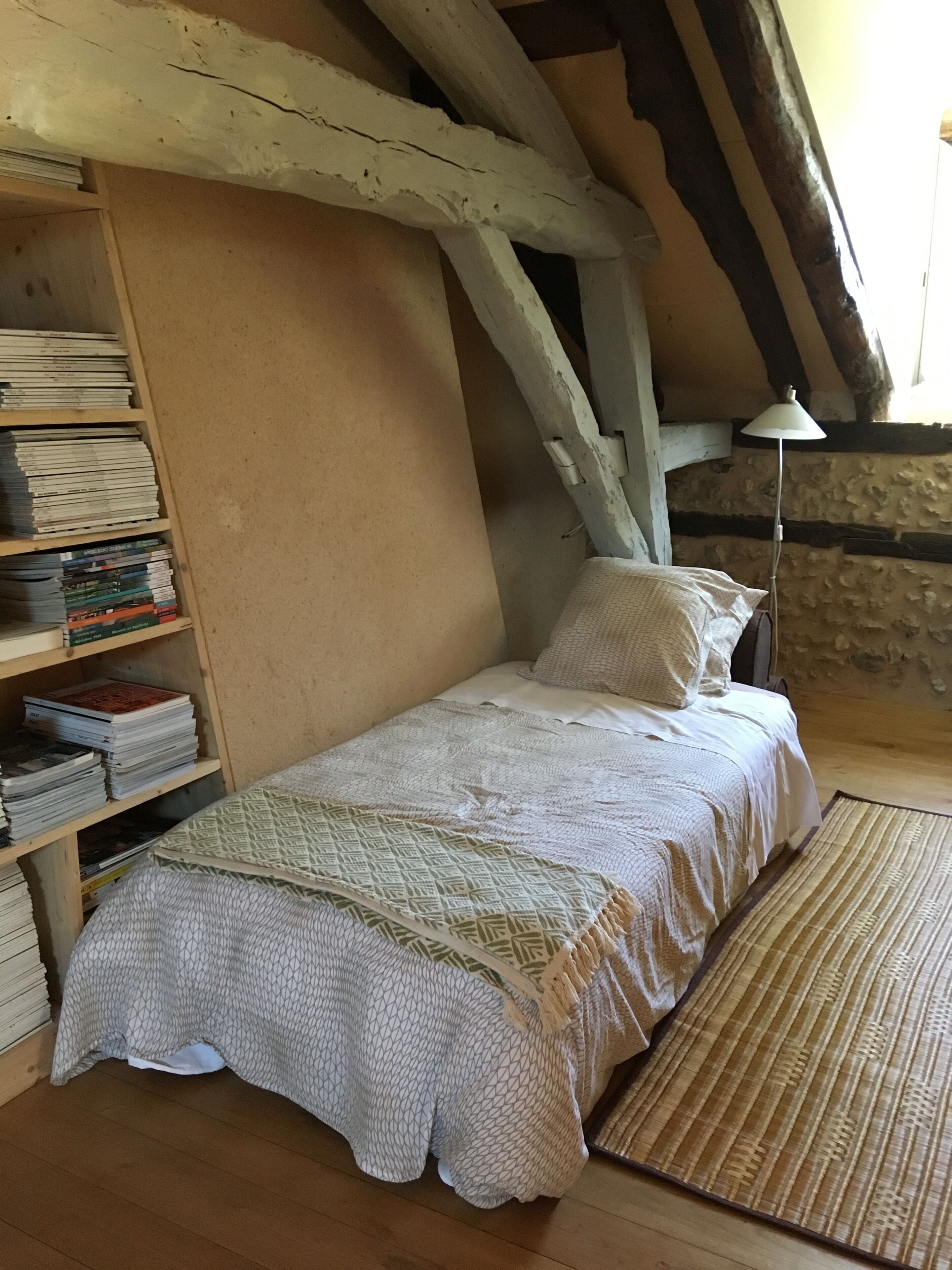 Ein Einzelbett mit einem gemusterten Überwurf, ein hölzernes Bücherregal voller Bücher und eine Steinwand.