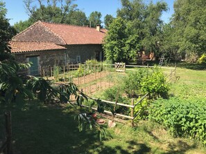 Una casa de piedra con techo de tejas, rodeada por un jardín vallado con diversas plantas y una cerca de madera.
