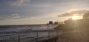 Eine Holzstege, der sich ins Meer erstreckt, mit Metallgeländer, einem Rettungsschwimmturm und einem Sonnenuntergang im Hintergrund.
