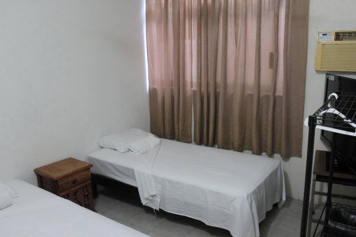 A small, clean room with two single beds, a wooden nightstand, a desk with a chair, and a window with curtains.