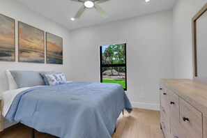 Een slaapkamer met een bed, een dressoir, een plafondventilator en een raam met uitzicht op groen.