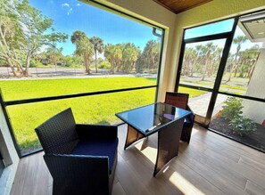 Een kamer met een glazen tafel, twee stoelen, grote ramen en uitzicht op een grasveld met bomen.