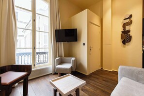 A living room with a sofa, a chair, a small table, a TV mounted on the wall, and a window with curtains. - JUST MOMENTS FROM LA MADELEINE, THE ICONIC FAUBOURG SAINT-HONORÉ (Paris)