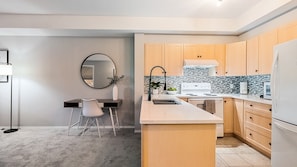 A modern kitchen with a central island, wooden cabinets, a white refrigerator, and a dining area with a chair and a round mirror. - Mountain View Condo | Pool, Hot Tub & Golf Nearby 1207 (Kelowna)