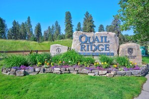 A sign for Quail Ridge Community with a stone border and flower bed. - Mountain View Condo | Pool, Hot Tub & Golf Nearby 1207 (Kelowna)