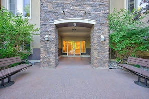 A stone entrance with benches, a glass door, and a building with a beige facade. - Mountain View Condo | Pool, Hot Tub & Golf Nearby 1207 (Kelowna)