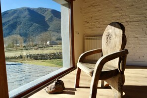 A wooden chair near a window with a view of mountains, a brick wall, and a tiled floor.